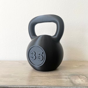 May include: A black kettlebell with a rounded body and a handle. The number "35" is embossed on the front within a circular frame. The kettlebell is made of a solid, matte material and sits on a wooden surface.
