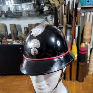 May include: A black military helmet with a red trim and a silver emblem. The helmet is displayed on a white mannequin head. The number "372" is visible on a white label. The background includes military artifacts.