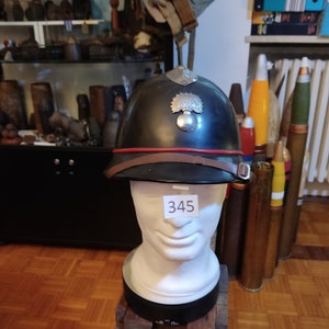 Può includere: Elmetto militare nero con una fascia rossa e un emblema argentato raffigurante una granata e un albero stilizzato. L'elmetto è su una testa di manichino bianca con un'etichetta che recita "345".