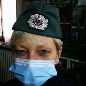 May include: A woman wearing a green military-style cap with a silver emblem and a blue surgical mask. The cap has a red and white symbol on the emblem.