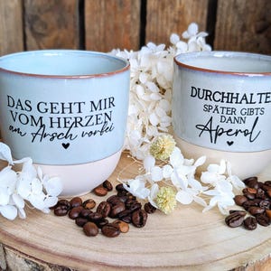 Könnte beinhalten: Zwei Keramikbecher mit hellblauen Oberteilen und cremefarbenen Böden. Auf einem Becher steht "DAS GEHT MIR VOM HERZEN am Arsch vorbei" und auf dem anderen "DURCHHALTEN! SPÄTER GIBTS dann Aperol". Die Becher sind von Kaffeebohnen und weißen Blumen umgeben.