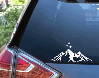 Könnte beinhalten: Ein weißer Vinyl-Aufkleber auf einer Heckscheibe, der die Silhouette eines stilisierten Gebirgszuges mit drei vierzackigen Sternen über dem zentralen Gipfel zeigt und ein klares, kontrastreiches grafisches Design auf dem Glas bildet.