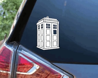 May include: A white vinyl decal of a classic British police box, featuring a rectangular structure with multiple window panes and a flat roof, applied to the rear window of a dark gray vehicle. The design is rendered in simple line art.