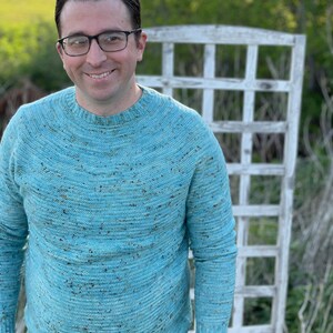 Peut inclure: Un pull bleu clair à col rond avec un motif moucheté. La personne qui porte le pull porte des lunettes. Un treillis blanc et de la verdure sont en arrière-plan.