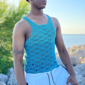 May include: A turquoise crochet tank top with a unique open weave design. The top features a scoop neck and wide straps. The garment is worn with white shorts. The background includes a blurred view of a body of water and greenery.