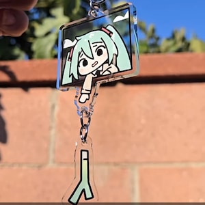 Pode incluir: Chaveiro de acrílico com um personagem de desenho animado com cabelo turquesa e expressão de surpresa. O chaveiro tem um design retangular com um fecho prateado e uma forma menor e estilizada abaixo. Os textos "ACRYLIC KEYCHAIN" e "DECALZOMBIE" são visíveis.