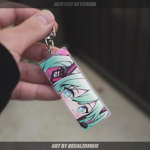 May include: A clear acrylic keychain with a colorful anime character design. The character has green hair, blue eyes, and is wearing a pink and black headset. The text "01" is visible on the headset. The keychain is labeled "Art by Decalzombie".