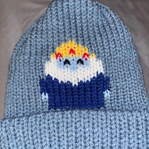 May include: A light blue knit beanie featuring a pixelated cartoon character. The character has a white face, blue body, and yellow and red accents. The beanie is made of a soft, ribbed knit material.