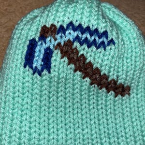 Op de afbeelding: Een lichtblauwe gebreide muts met een pixelachtig pikhouweelontwerp in bruin en blauw. De muts is gemaakt van zacht, getextureerd garen, ideaal om warm te blijven. Het pikhouweelontwerp is een opvallend detail.