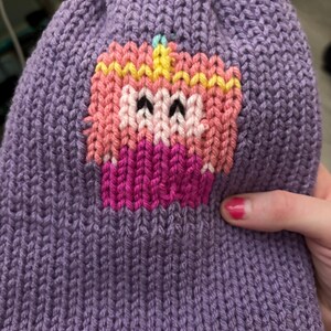Op de afbeelding: Een paarse gebreide muts met een cupcake-motief. De cupcake is geborduurd met roze, gele en witte draden. De muts is gemaakt van een zacht, getextureerd materiaal. Het cupcake-ontwerp bevindt zich aan de voorkant van de muts.