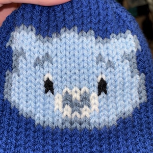 May include: A blue knitted hat featuring a light blue and white cartoon monster face. The hat has black eyes and a white mouth. The hat is made of a soft, textured yarn, perfect for cold weather.