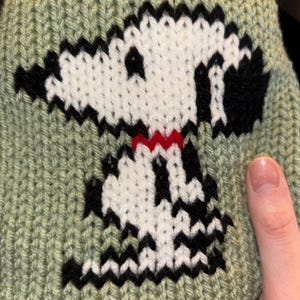 May include: A sage green knit hat featuring a white, black, and red intarsia design of a cartoon dog. The dog is depicted in profile with a white body, black ears and nose, and a red collar, set against the textured vertical ribbed knit fabric.