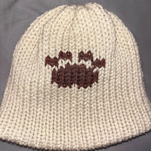 May include: Cream-colored knit beanie with a brown crab design. The hat is made of a thick, ribbed knit material. The crab design is centered on the front of the hat. The hat has a rounded crown and a slightly flared brim.