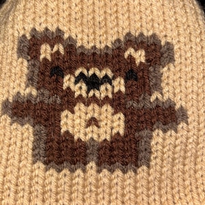 May include: A knitted beanie in a light beige color, featuring a brown and gray pixelated design of a bear-like character. The hat is made of a textured, ribbed knit fabric.