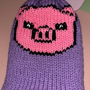 May include: Purple knitted beanie with a pink pig face design. The pig has black eyes and a black snout.