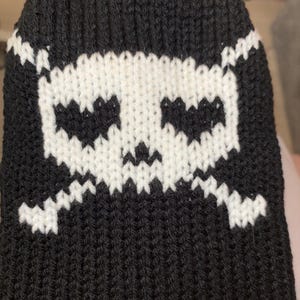May include: Black knit beanie featuring a white skull and crossbones design. The skull has two eye sockets and a jagged mouth. The beanie is made of a soft, ribbed knit material, perfect for cold weather.