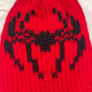 May include: A red knit beanie with a black geometric design. The hat is made of a thick, textured yarn and features a repeating pattern across the front. The beanie is a warm winter accessory.