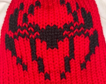 Puede incluir: Un gorro rojo tejido con un diseño geométrico negro. El gorro está hecho de un hilo grueso y texturizado y presenta un patrón repetitivo en la parte delantera. Un cálido accesorio de invierno.