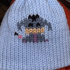 May include: A light blue knitted beanie hat with a pixelated cartoon monster design in grey, black, tan, and purple. A small pink bat is on the left side. The hat is made of a ribbed knit fabric.