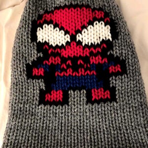 May include: Gray knit beanie featuring a pixelated Spider-Man design. The superhero's face and body are rendered in red, white, blue, and black against the gray background. The hat is made of a thick, textured knit.