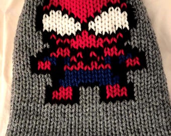 Puede incluir: Gorro de punto gris con un diseño pixelado de Spider-Man. La cara y el cuerpo del superhéroe están representados en rojo, blanco, azul y negro sobre un fondo gris. El gorro está hecho de un punto grueso y texturizado.