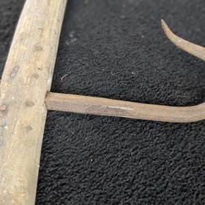 Könnte beinhalten: Nahaufnahme eines Vintage-Fleischhakens mit Holzgriff. Der Haken besteht aus rostigem Metall und ist mit einem Metallverschluss am Holzgriff befestigt. Das Holz ist verwittert und weist Altersspuren auf.