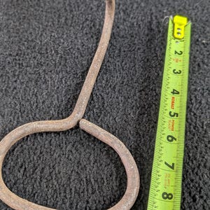 Könnte beinhalten: Ein rostiges Metallwerkzeug mit einem geschlungenen Griff und einem gebogenen, spitzen Ende. Ein gelbes und grünes Maßband liegt neben dem Werkzeug und zeigt die Maße in Zentimetern an.