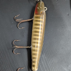 May include: Vintage fishing lure with a metallic gold and brown body, featuring a scale pattern. The lure has three treble hooks and a red accent near the head. A metal attachment is at the top.
