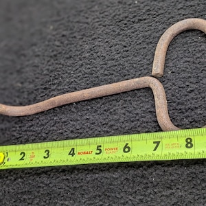 Könnte beinhalten: Ein rostiger Metallhaken mit gebogenem Ende und Schlaufengriff. Der Haken ist etwa 18 cm lang, gemessen mit einem grün-gelben Maßband. Das Objekt ist wahrscheinlich ein antikes Werkzeug.