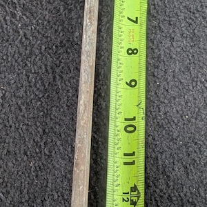 Könnte beinhalten: Ein rostiger Metallhaken mit gebogenem Ende, neben einem grün-gelben Maßband. Das Band zeigt Messungen bis zu 40 cm. Der Haken ist wahrscheinlich ein Werkzeug zum Heben oder Ziehen.