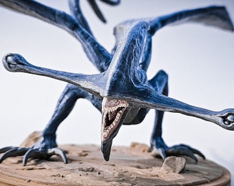 May include: A detailed blue sculpture of a creature with large wings and a long beak, posed on a circular base. The sculpture has a detailed texture and is painted in shades of blue. The base is a light brown color, with a sandy texture.