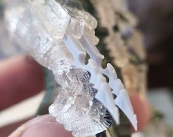 May include: A detailed figurine of a creature with a camouflage pattern in shades of green and brown. The figure has sharp, clear, claw-like structures extending from its hand. The figure is held by a person.