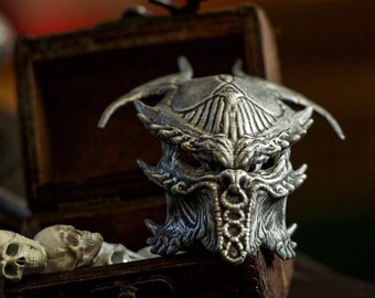 May include: A detailed silver-colored mask with an intricate design, resembling a creature's face, sits atop a small wooden chest. The chest is open, revealing small skulls and a decorative item. The background is blurred, with a logo in the bottom right corner.