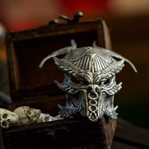May include: A detailed silver-colored mask with an intricate design, resembling a creature's face, sits atop a small wooden chest. The chest is open, revealing small skulls and a decorative item. The background is blurred, with a logo in the bottom right corner.