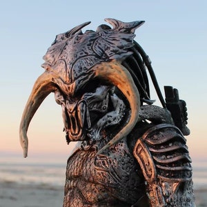 May include: A detailed Predator action figure, featuring a silver and bronze color scheme. The figure has intricate armor, a horned helmet, and a spear. The background shows a beach with a sunset.