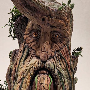 May include: A detailed sculpture of a tree-like figure with a long beard and a face carved into the wood. The sculpture is painted in shades of brown, green, and gray, with small green leaves and moss accents. The base is gray.