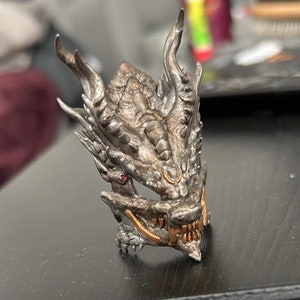 May include: A detailed, silver-toned dragon head sculpture with intricate scales, horns, and a menacing expression. The dragon's mouth is open, revealing sharp teeth with gold accents. A small red gem is set in the eye.