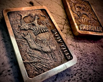 May include: Two rectangular metal bars with detailed skull and skeletal hand designs. One bar reads "STILL ALIVE" and the other reads "UNBROKEN". The bars have a bronze-like color with silver edges.
