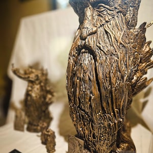 May include: A detailed bronze-colored sculpture of a tree-like figure with a textured surface. The sculpture has a beard and a stern expression. A smaller version of the sculpture is in the background. The sculpture is on a white surface.