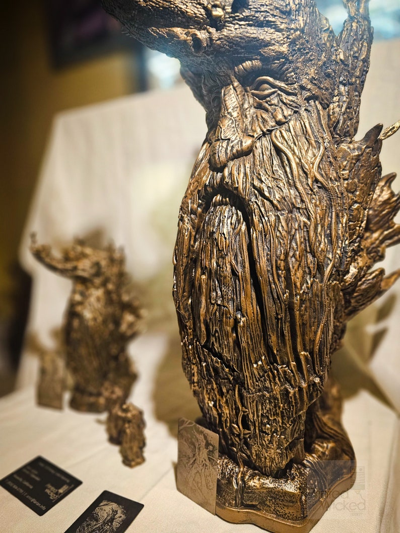 May include: A detailed bronze-colored sculpture of a tree-like figure with a textured surface. The sculpture has a beard and a stern expression. A smaller version of the sculpture is in the background. The sculpture is on a white surface.
