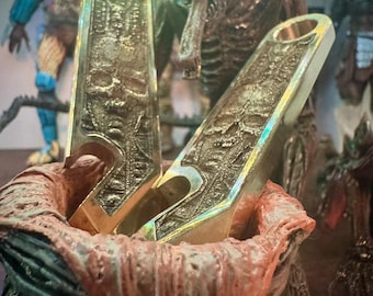 May include: Two gold-colored, rectangular pendants with skull and intricate designs are displayed in a red and green container. The pendants have a hole at the top. The background features Predator and Alien figures.