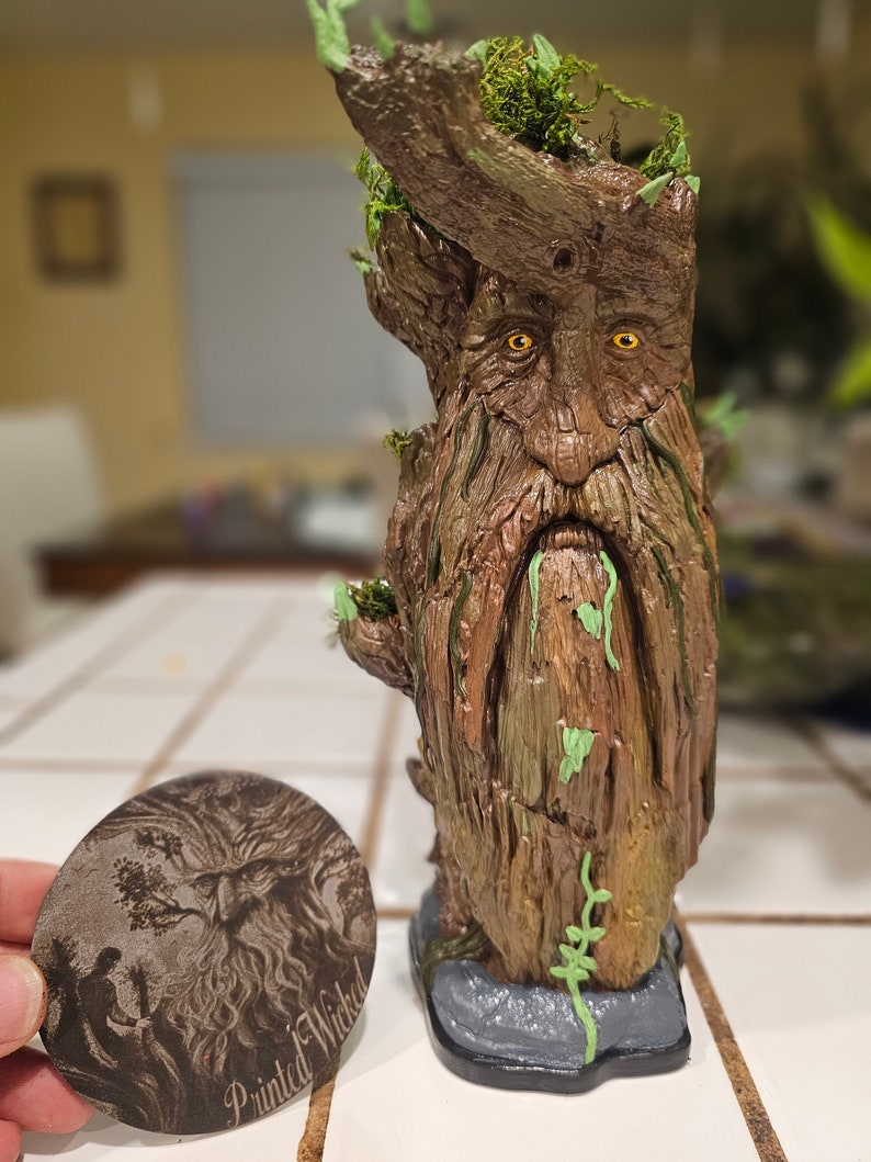 May include: A detailed, hand-painted tree-like sculpture with a face, beard, and moss accents. The sculpture has a weathered brown finish with green highlights. A wooden, oval-shaped item with the text "Painted Wicked" and a detailed illustration is next to the sculpture.