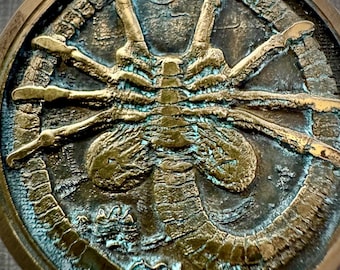 May include: A circular, bronze-colored metal object with a detailed relief of a creature. The design features a central, elongated body with multiple limbs and a textured surface. The outer rim has a decorative pattern.