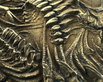 May include: Close-up of a detailed, bronze-colored relief sculpture. The artwork depicts a skull with intricate textures and shadows, suggesting a gothic or fantasy theme. The relief has a weathered appearance, enhancing the aged aesthetic.