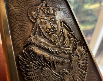 May include: A rectangular, bronze-colored metal object with an engraved image of a man wearing a crown and elaborate clothing. The man has a long beard and is flanked by two eagles. The background features a wave-like pattern.
