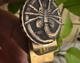 May include: A brass money clip featuring a detailed, circular design of an insect-like creature. The clip has a gold finish and a hinged design for securing bills. The design is in black and gold.