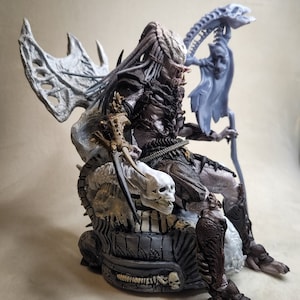 May include: A detailed Predator action figure seated on a throne of skulls and bones. The figure is primarily gray, brown, and black, with intricate armor and dreadlocks. The throne has a skeletal design, and the figure holds a weapon.