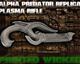 May include: A silver Alpha Predator Replica Plasma Rifle, with intricate, organic-looking details. The text "ALPHA PREDATOR REPLICA PLASMA RIFLE" is at the top, and "PRINTED WICKED" is at the bottom. The background has a textured, metallic appearance.