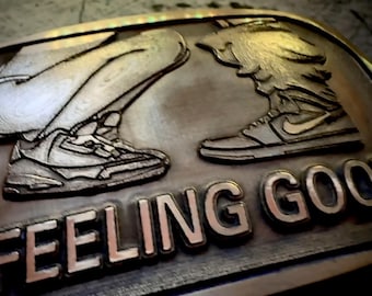 May include: A silver-colored metal belt buckle with the words "FEELING GOOD" in raised letters. The buckle features detailed depictions of two sneakers, one on each side, suggesting a theme of athletic footwear and well-being.