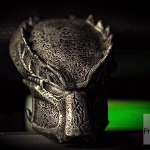Op de afbeelding: Een gedetailleerde, metalen Predator-masker sculptuur. De sculptuur is een donker, getextureerd zilver met ingewikkelde details, waaronder de kenmerkende kaken en dreadlocks van de Predator. Het masker staat tegen een donkere achtergrond met een groene gloed.
